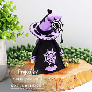 Back view of handmade 3D felt Fergus the Gnome created with Spellbinders dies by Sandi MacIver. The gnome wears a purple hat with black bat accents and a dangling spiderweb, along with a black robe decorated with purple spiderweb and bat embellishments. Displayed on a wooden slice with greenery in the background.