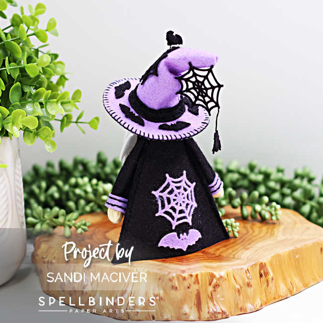 Back view of handmade 3D felt Fergus the Gnome created with Spellbinders dies by Sandi MacIver. The gnome wears a purple hat with black bat accents and a dangling spiderweb, along with a black robe decorated with purple spiderweb and bat embellishments. Displayed on a wooden slice with greenery in the background.