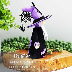 ide view of handmade 3D felt Fergus the Gnome created with Spellbinders dies by Sandi MacIver. The gnome wears a purple hat with a stitched black spiderweb and dangling felt spider, a black robe with purple accents, and bat embellishments. Displayed on a wooden slice with greenery in the background.