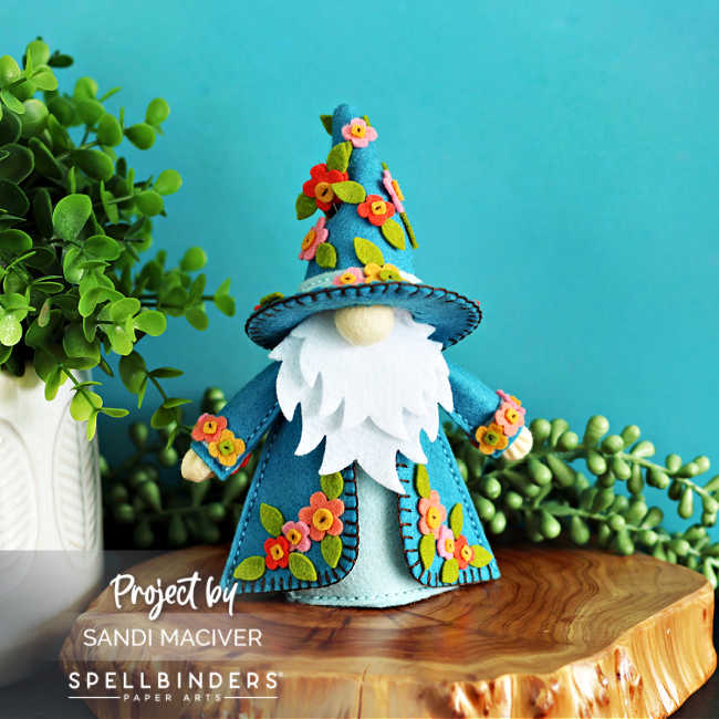 Handmade 3D felt Fergus the Gnome created with Spellbinders dies by Sandi MacIver. This version features a teal coat and hat decorated with bright stitched felt flowers in shades of pink, orange, and yellow with green leaves. Displayed on a wooden slice with greenery and a turquoise background.
