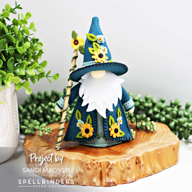 Handmade 3D felt Fergus the Gnome created with Spellbinders dies by Sandi MacIver. The gnome wears a deep teal robe and tall hat decorated with yellow sunflowers and green leaves, holding a gold-striped staff. Displayed on a wooden slice with greenery in the background.