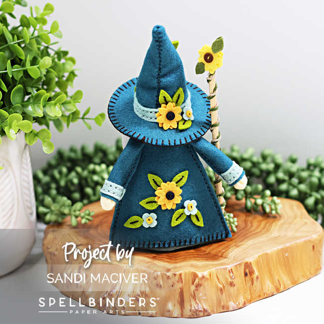 Back view of handmade 3D felt Fergus the Gnome created with Spellbinders dies by Sandi MacIver. The gnome wears a teal robe and tall hat, both decorated with stitched yellow sunflowers and green leaves, and holds a sunflower-topped staff. Displayed on a wooden slice with greenery in the background.