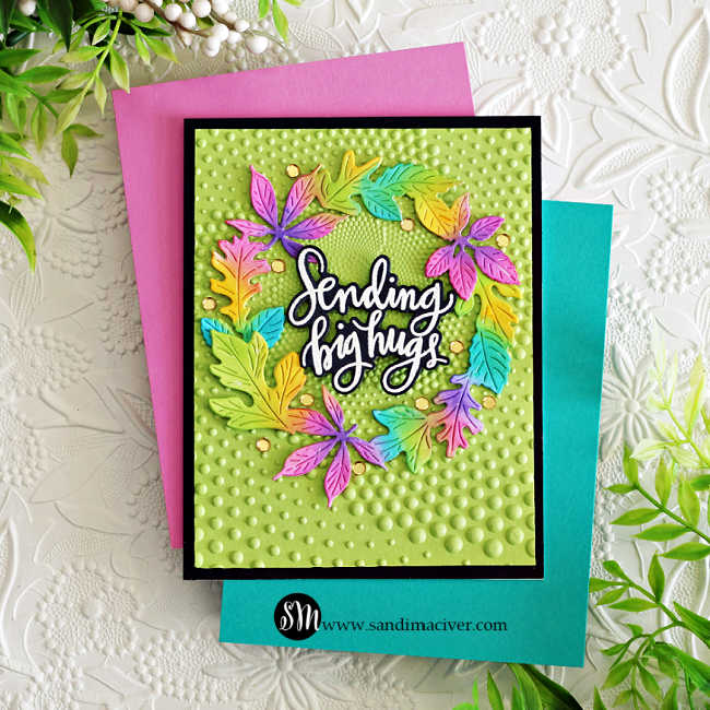 Handmade greeting card featuring a colorful die-cut fall wreath with leaves in bright shades of green, pink, purple, yellow, and turquoise. The wreath frames a white script sentiment that reads “Sending big hugs.” The background is embossed with a dotted spiral pattern on light green cardstock, mounted on a black mat and placed over a pink and teal background for display.