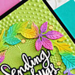 Handmade greeting card featuring a colorful die-cut fall wreath with leaves in bright shades of green, pink, purple, yellow, and turquoise. The wreath frames a white script sentiment that reads “Sending big hugs.” The background is embossed with a dotted spiral pattern on light green cardstock, mounted on a black mat and placed over a pink and teal background for display.