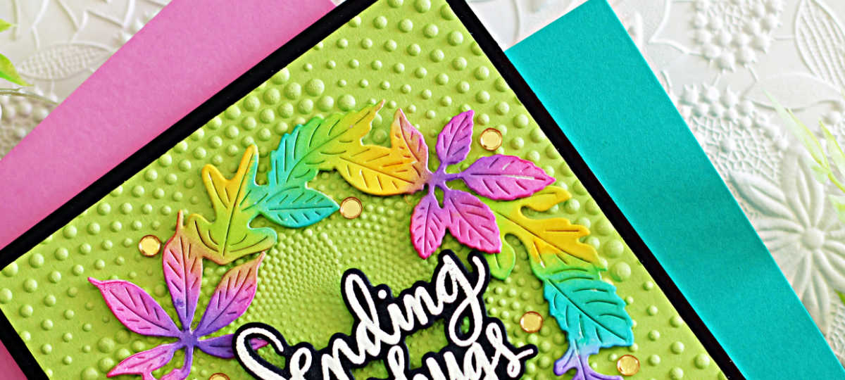 Handmade greeting card featuring a colorful die-cut fall wreath with leaves in bright shades of green, pink, purple, yellow, and turquoise. The wreath frames a white script sentiment that reads “Sending big hugs.” The background is embossed with a dotted spiral pattern on light green cardstock, mounted on a black mat and placed over a pink and teal background for display.