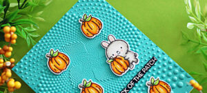 Handmade card featuring stamped and die-cut images from the Simon Says Stamp Pumpkin Pals set, colored with Copic markers. The design includes a cute gray bunny hugging an orange pumpkin, surrounded by additional pumpkins with green leaves. The background is embossed with the Dotted Spiral embossing folder on bright turquoise cardstock, adding texture and dimension.