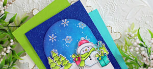 Handmade holiday card featuring the Simon Says Stamp and Memory Box Good Cheer collaboration stamp and die set. The design showcases an ink blended arch with a cosmic night sky, layered over a deep blue snowflake embossed background. A cheerful snowman in a pink hat and green scarf is surrounded by colorful presents, with bright green trees and snowflakes adding festive detail. Sentiment reads “Holiday Hugs.”