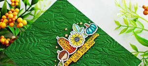 Handmade fall-themed greeting card created with the Spellbinders September Clear Stamp, Die, and Embossing Folder Clubs. The background features a rich green embossed panel with a leafy vine pattern. At the center, there’s a cluster of colorful stamped and die-cut images including a sunflower, acorn, gourd, and teal pumpkin, along with a sentiment strip reading “A LITTLE FALL CHARM, JUST FOR YOU.” Small golden gems accent the design, adding sparkle.