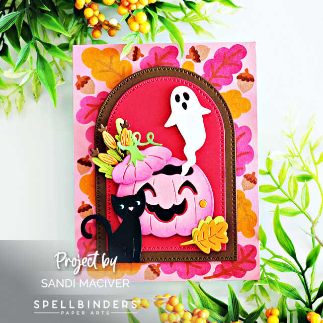 Handmade Halloween card created with the Spellbinders September Small Die of the Month, Stencil of the Month, and Perfect Touch Arches and Tags dies. The background features a pink panel stenciled with layered autumn leaves in orange, gold, and magenta. At the center, a stitched arch frame in deep red and brown showcases a smiling pink jack-o’-lantern topped with flowers and leaves, accented by a playful black cat and a friendly white ghost. A yellow die-cut leaf embellishment completes the festive fall design.