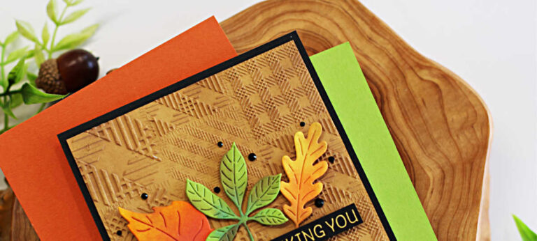 Handmade fall-themed thank you card created with the Spellbinders September 3D Embossing Folder and Large Die of the Month kits. The background is embossed with a woven plaid texture in kraft cardstock, layered on black cardstock. Three colorful die-cut leaves in orange, green, and yellow are arranged at the center. A black sentiment strip with gold embossing reads “THIS IS ME THANKING YOU.” The card is displayed on a wood slice with coordinating orange and green envelopes, surrounded by fall foliage and acorns.