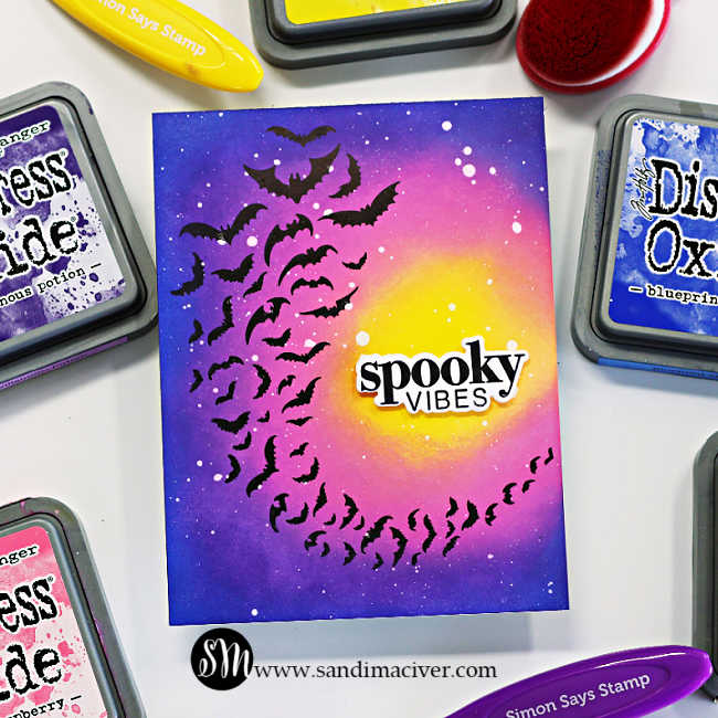 Handmade Halloween card featuring a bold ink-blended background created with Distress Oxide inks. The design showcases a swarm of black bats spiraling across a glowing yellow moon, fading into pink and purple night sky tones. White paint splatters add a starry effect, and the sentiment “spooky vibes” is die cut and layered in black and white at the center. Card designed by Sandi MacIver using the Simon Says Stamp Halloween Basics Stamp and Die Set.