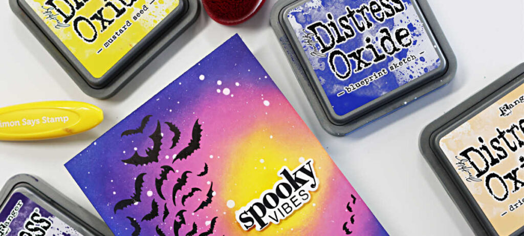 Handmade Halloween card featuring a bold ink-blended background created with Distress Oxide inks. The design showcases a swarm of black bats spiraling across a glowing yellow moon, fading into pink and purple night sky tones. White paint splatters add a starry effect, and the sentiment “spooky vibes” is die cut and layered in black and white at the center. Card designed by Sandi MacIver using the Simon Says Stamp Halloween Basics Stamp and Die Set.