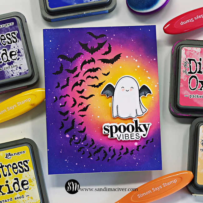 Handmade Halloween card featuring a glowing Distress Oxide ink–blended background in yellow, pink, and purple tones. A swirl of black bats frames a cute white ghost with black bat wings and a smiling face. The sentiment “spooky vibes” is die cut in black and white beneath the ghost. Card created by Sandi MacIver using the Simon Says Stamp Halloween Basics and My Boo stamp and die sets.