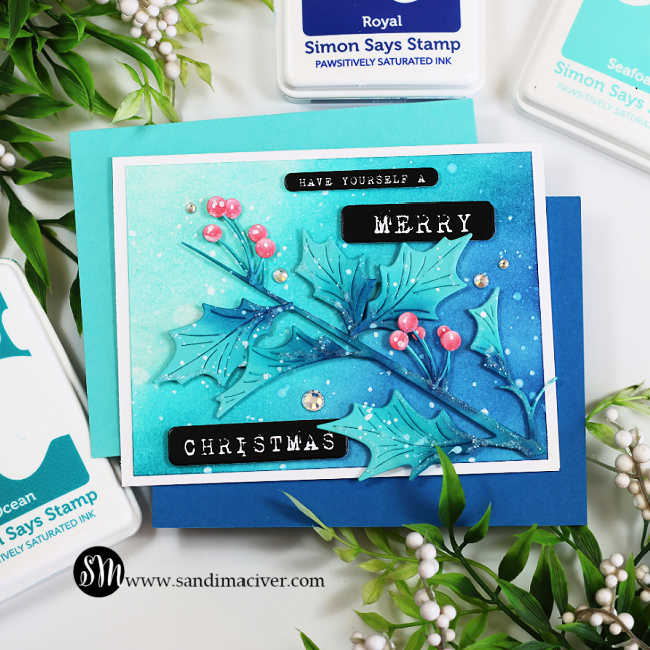 Handmade Christmas card featuring the Simon Says Stamp Narrow Holly Cluster Die Set, ink blended in Pawsitively Saturated Inks in shades of Ocean, Seafoam, Royal, and Rhubarb. The holly leaves and berries are accented with white paint splatter and gem embellishments on a softly blended teal-to-blue background. The sentiment reads “Have Yourself a Merry Christmas” in white typewriter-style font on black labels, layered over a white card base with coordinating blue and teal mats. Designed by Sandi MacIver for a quick and easy holiday card.
