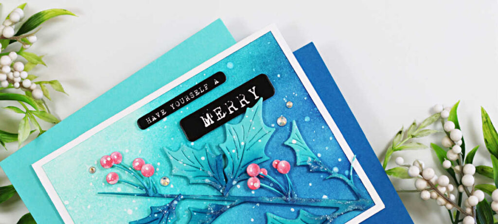 Handmade Christmas card featuring the Simon Says Stamp Narrow Holly Cluster Die Set, ink blended in Pawsitively Saturated Inks in shades of Ocean, Seafoam, Royal, and Rhubarb. The holly leaves and berries are accented with white paint splatter and gem embellishments on a softly blended teal-to-blue background. The sentiment reads “Have Yourself a Merry Christmas” in white typewriter-style font on black labels, layered over a white card base with coordinating blue and teal mats. Designed by Sandi MacIver for a quick and easy holiday card.