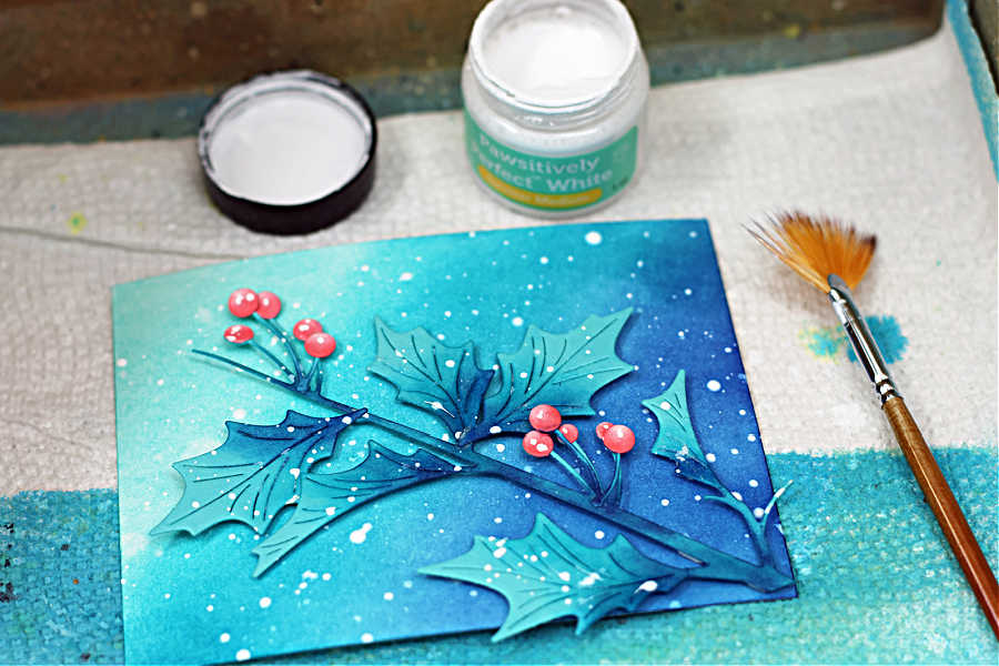 Handmade Christmas card panel featuring die-cut holly leaves and berries from the Simon Says Stamp Narrow Holly Cluster Die Set. The background, ink blended in Pawsitively Saturated Inks in Ocean, Seafoam, and Royal, has been lightly spritzed with water for texture and **splattered with white paint