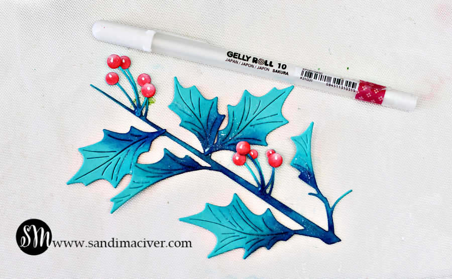Close-up of die-cut holly leaves and pink berries from the Simon Says Stamp Narrow Holly Cluster Die Set, ink blended in Ocean, Royal, and Rhubarb Pawsitively Saturated Inks. A Sakura Gelly Roll #10 white pen is used to add small white highlight dots to the tops of the berries for added shine and dimension. Designed by Sandi MacIver for Simon Says Stamp