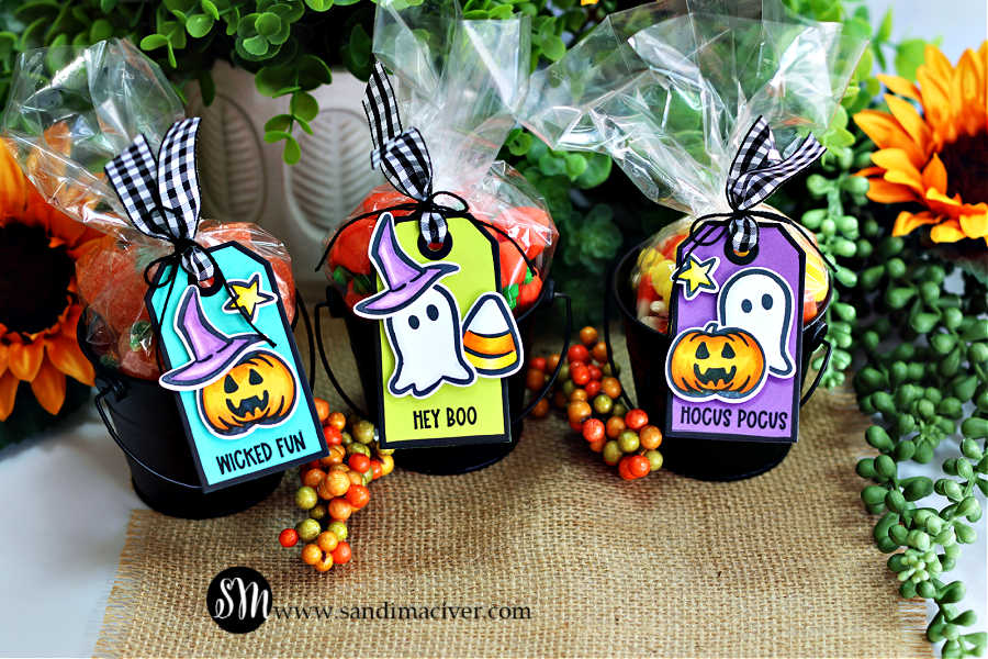 Three black metal pails filled with colorful Halloween candies are displayed on a burlap surface, each topped with a clear cello bag tied with black-and-white gingham ribbon. Attached to each pail is a bright handmade tag created with the Tim Holtz Tiny Frights Stamp Set by Simon Says Stamp. The tags feature playful Halloween images — pumpkins, ghosts, witch hats, stars, and candy corn — in vibrant teal, lime, and purple backgrounds with fun sentiments that read “Wicked Fun,” “Hey Boo,” and “Hocus Pocus.” Sunflowers and greenery frame the festive scene.