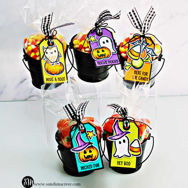Five black metal pails filled with colorful Halloween candies are topped with clear treat bags tied with black-and-white gingham ribbon. Each pail features a bright handmade tag created with the Tim Holtz Tiny Frights Stamp Set, showcasing cute Halloween images—a ghost, owl, pumpkin, bat, and candy—colored in vibrant shades of orange, teal, purple, and lime green. The tags display playful sentiments like “What a Hoot,” “Wicked Fun,” “Here for the Candy,” “Hey Boo,” and “Hocus Pocus,” making them perfect for festive Halloween gift giving.
