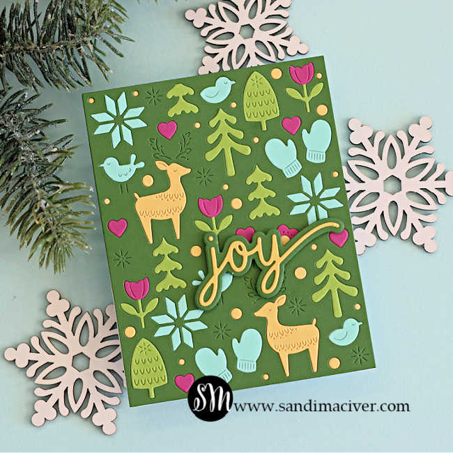 Handmade holiday card created with the Simon Says Stamp Wintertime Celebration Plate featuring a green background with die-cut winter icons—reindeer, trees, mittens, hearts, snowflakes, and birds—in cheerful pastel colors and a layered ‘joy’ sentiment in gold and aqua.