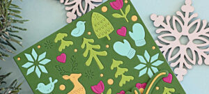 Handmade holiday card created with the Simon Says Stamp Wintertime Celebration Plate featuring a green background with die-cut winter icons—reindeer, trees, mittens, hearts, snowflakes, and birds—in cheerful pastel colors and a layered ‘joy’ sentiment in gold and aqua.