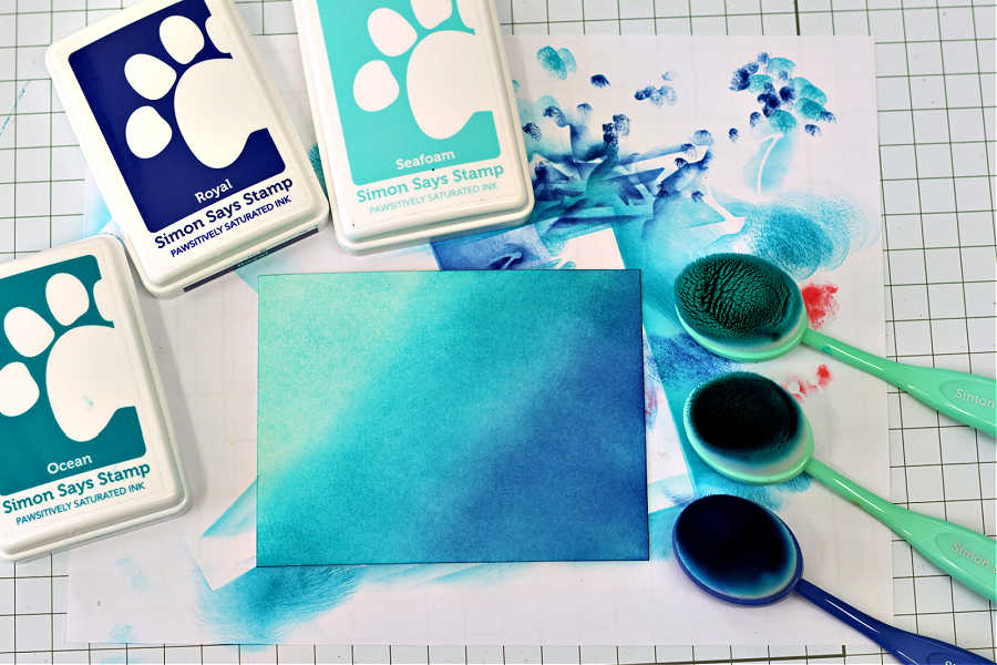 nk blending background for a handmade Christmas card using Simon Says Stamp Pawsitively Saturated Inks in Ocean, Seafoam, and Royal. The inks are blended smoothly onto a 4" x 5-1/4" piece of watercolor cardstock using coordinating Simon Says Stamp blending brushes. The workspace shows vibrant blue and teal tones with ink pads and brushes arranged around the blended panel. Designed by Sandi MacIver for Simon Says Stamp.