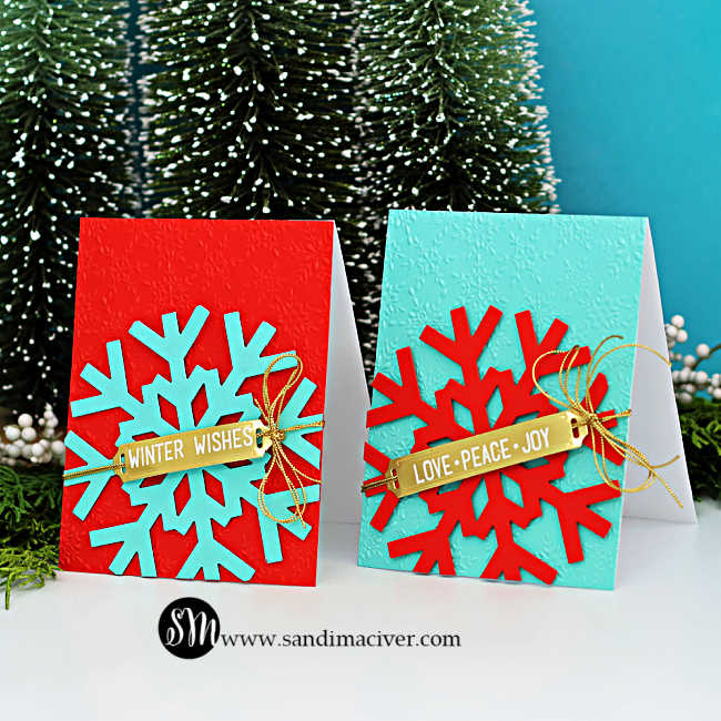 wo handmade Christmas cards featuring the Spellbinders Large Snowflake Die in bold color combos of Poppy and Waterfall cardstock. Each card has an embossed background—one in red with a teal snowflake and “Winter Wishes” gold label, and the other in teal with a red snowflake and “Love • Peace • Joy” gold label. Both cards are finished with shiny gold cord bows for a festive, modern holiday look.
