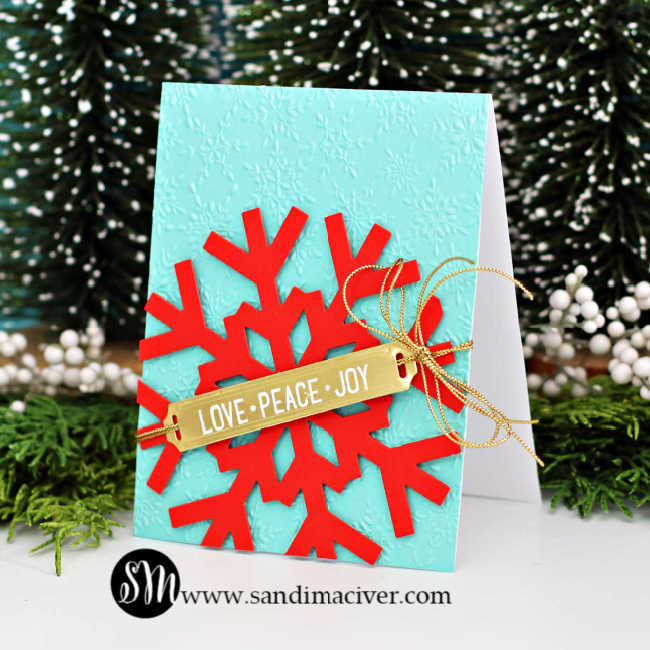 A handmade Christmas card featuring a large red snowflake die cut from Spellbinders Poppy cardstock layered over an embossed panel of Spellbinders Waterfall cardstock. The card is embellished with a gold “LOVE • PEACE • JOY” label from the Label Slider stamp and die set, tied with shiny gold cord from Simon Says Stamp. The festive color combo pops beautifully against a backdrop of snowy evergreens.