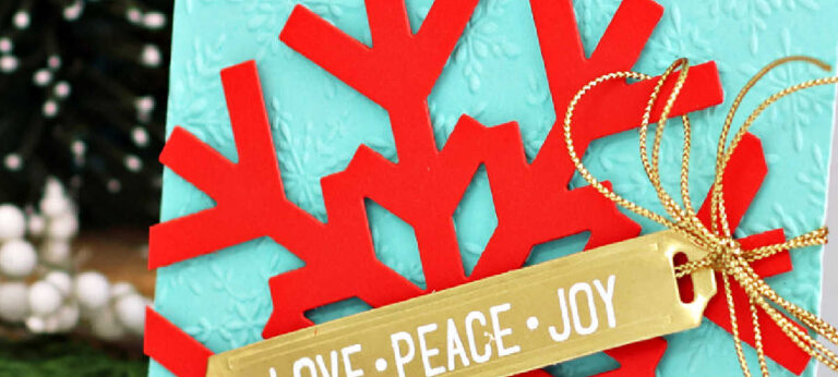 A handmade Christmas card featuring a large red snowflake die cut from Spellbinders Poppy cardstock layered over an embossed panel of Spellbinders Waterfall cardstock. The card is embellished with a gold “LOVE • PEACE • JOY” label from the Label Slider stamp and die set, tied with shiny gold cord from Simon Says Stamp. The festive color combo pops beautifully against a backdrop of snowy evergreens.