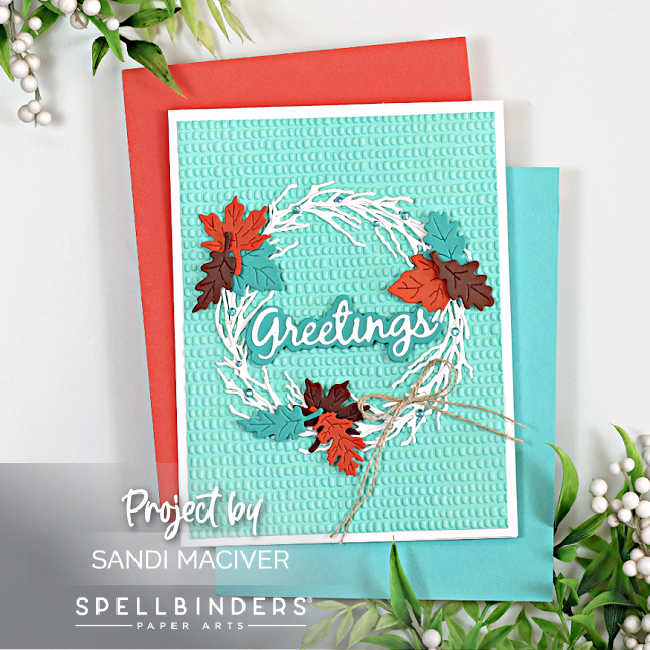 Handmade fall greeting card created with the Spellbinders Autumn Greetings Wreath Die Set and Dimpled 3D Embossing Folder. The card features a white twig wreath adorned with die-cut maple leaves in rich autumn colors—rust, teal, and chocolate brown. The sentiment “Greetings” appears in white with a teal shadow layer across the center of the wreath, accented by a small natural twine bow. The background is embossed with a raised dimple texture on aqua cardstock, mounted on a white card base, and displayed with red and teal cardstock mats surrounded by greenery and white berries.