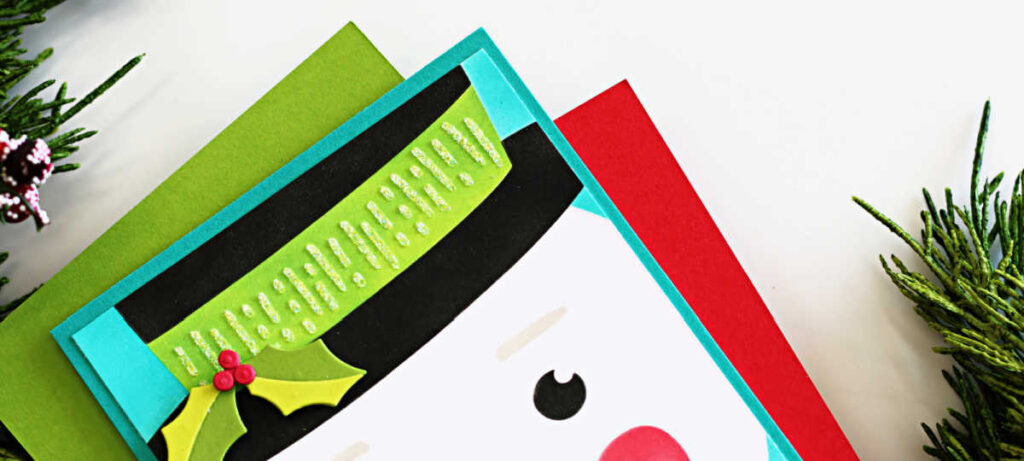 Handmade Frosty snowman Christmas card created with the Spellbinders Stencil of the Month Club ‘Frosty Wishes.’ Features a cheerful snowman face with a carrot nose, rosy cheeks, and a green top hat accented with holly, layered on a bright turquoise card base for a fun and festive holiday design.