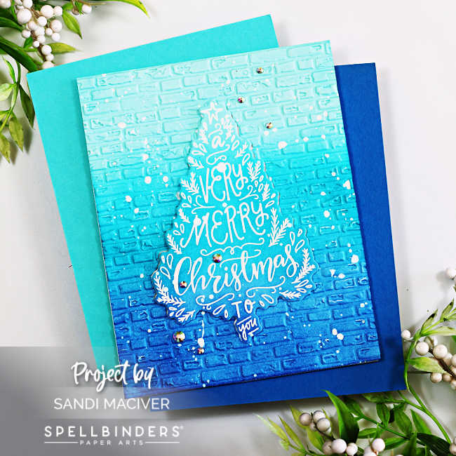 Handmade holiday card created by Sandi MacIver using the Spellbinders To You and Yours Clear Stamp and Die of the Month and the 3D Embossing Folder of the Month. The design features a blue ombré ink-blended background embossed with a brick pattern, overlaid with a white embossed Christmas tree-shaped sentiment that reads “A Very Merry Christmas to You.” The card is accented with white paint splatters and small clear gems for sparkle, layered over teal and navy blue cardstock panels.