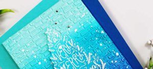 Handmade holiday card created by Sandi MacIver using the Spellbinders To You and Yours Clear Stamp and Die of the Month and the 3D Embossing Folder of the Month. The design features a blue ombré ink-blended background embossed with a brick pattern, overlaid with a white embossed Christmas tree-shaped sentiment that reads “A Very Merry Christmas to You.” The card is accented with white paint splatters and small clear gems for sparkle, layered over teal and navy blue cardstock panels.