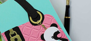 A handmade greeting card designed to look like a pink quilted purse with a teal blue background. The purse features a black strap with gold hardware and a cute panda bear charm hanging from a small gold chain. A gold and black sentiment reads “Charmed to Know You.” The card is surrounded by a fountain pen and greenery for styling. The bottom corner includes the text “Project by Sandi MacIver | Spellbinders Paper Arts.”