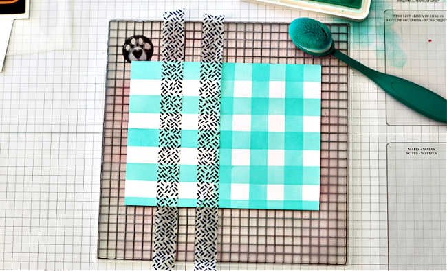 Photo showing how to create homemade gingham paper using Washi Tape and a Craft Tacky Mat. Strips of patterned washi tape are placed vertically over a turquoise and white ink-blended background, creating a checkered gingham effect. The Craft Tacky Mat’s built-in grid lines help align the tape evenly, while a teal blending brush and ink pad are visible to the side, demonstrating the ink-blending process.