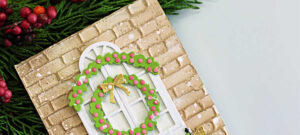 Handmade die-cut Christmas card with a white arched window, green wreath, and garland with gold bows against a brick embossed background, created using Simon Says Stamp dies