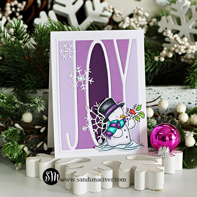 Handmade Christmas card featuring the Simon Says Stamp Joy Cover Die layered over a purple ombré background. White Harmony Snowflakes with gem centers accent the design, and a Copic-colored snowman from the Cozy in the Cold set stands at the bottom right holding a holly sprig with a red bird. The card is displayed on a bed of frosted greenery with the sandimaciver.com logo at the bottom.