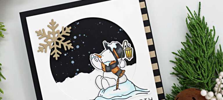 Handmade winter card featuring an illustrated snowman from the Simon Says Stamp Cozy in the Cold stamp and die set. The snowman sits on a snowbank holding a lantern and looking up at a dark night sky, isplattered with white paint to resemble falling snow. A kraft die-cut snowflake embellishes the top left corner, and the sentiment “Peace on Earth” is stamped below the scene. The card is framed with black and kraft striped paper and styled with greenery and rustic jingle bells for the photo.