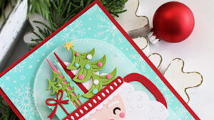 Handmade Christmas card featuring a die-cut Santa mug with a white beard, pink cheeks, and a red cup handle. The mug is filled with die-cut Christmas trees, a gift, and a star. The sentiment "Merry Christmas" appears in red at the bottom. The background is aqua cardstock with white glimmer-foiled snowflakes and stitched arch detailing, mounted on a red card base. Card is styled with festive greenery and berries in the background.