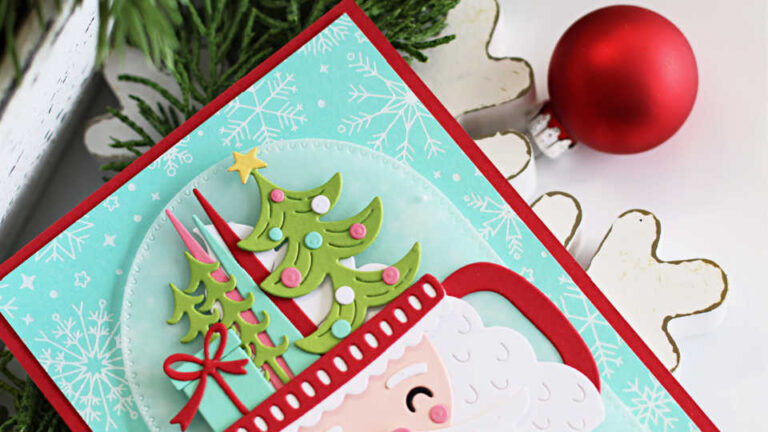 Handmade Christmas card featuring a die-cut Santa mug with a white beard, pink cheeks, and a red cup handle. The mug is filled with die-cut Christmas trees, a gift, and a star. The sentiment "Merry Christmas" appears in red at the bottom. The background is aqua cardstock with white glimmer-foiled snowflakes and stitched arch detailing, mounted on a red card base. Card is styled with festive greenery and berries in the background.