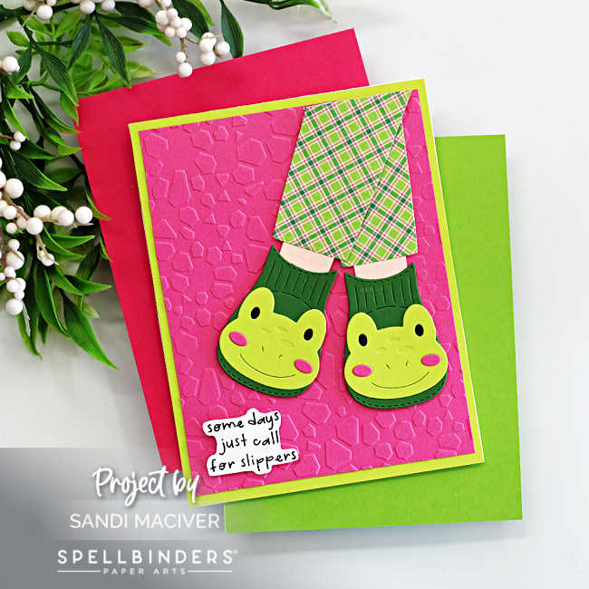 Handmade frog slipper card created with the Spellbinders Cozy Toes Friends Die Set and embossed with the Faceted Petals Embossing Folder. The card features cheerful green frog slippers paired with plaid pink-and-green pajama pants layered over a bright pink embossed background and a lime green card base. A playful sentiment strip reads “some days just call for slippers.” Designed by Sandi MacIver for Spellbinders Paper Arts.