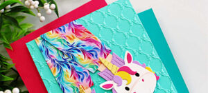 Handmade unicorn slipper card created with the Spellbinders Cozy Toes Friends Die Set and embossed using the Petite Bows 3D Embossing Folder. The card features colorful rainbow pajama pants tucked into whimsical unicorn slippers layered over an aqua embossed background with a playful sentiment strip that reads “I’d leave the house for you.” Designed by Sandi MacIver for Spellbinders Paper Arts.