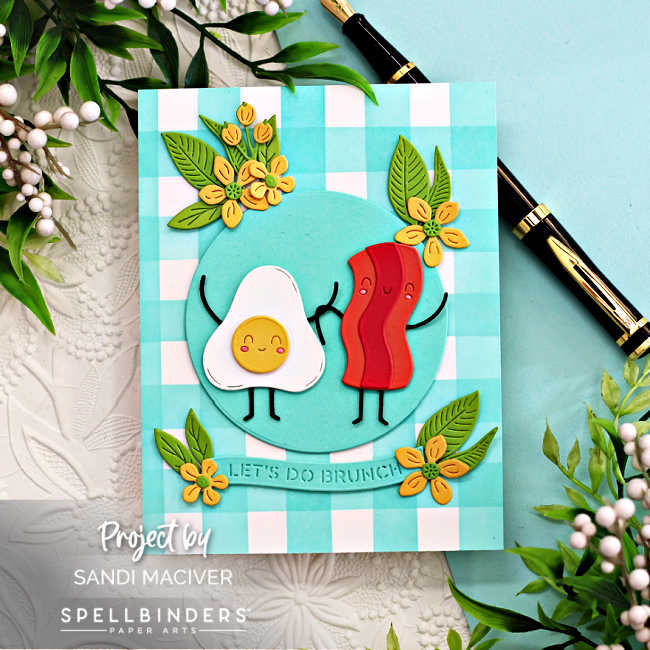 Handmade “Let’s Do Brunch” card created with the Spellbinders Small Die of the Month for November featuring a cute bacon and egg duo holding hands on a turquoise dinner plate. The card includes cheerful yellow flowers and green leaves die-cut with the Citrus Spritz Die Set, arranged around the plate. The background showcases a teal-and-white gingham pattern, ink blended with washi tape masking for the stripes. The design is bright, playful, and perfect for brunch-themed or friendship cards.