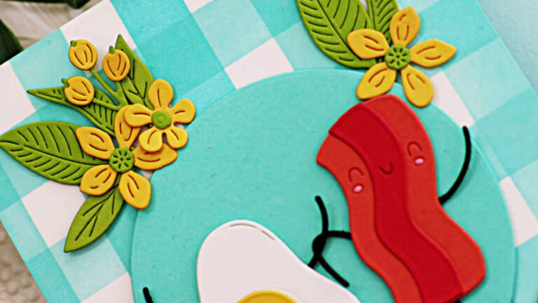 Handmade “Let’s Do Brunch” card created with the Spellbinders Small Die of the Month for November featuring a cute bacon and egg duo holding hands on a turquoise dinner plate. The card includes cheerful yellow flowers and green leaves die-cut with the Citrus Spritz Die Set, arranged around the plate. The background showcases a teal-and-white gingham pattern, ink blended with washi tape masking for the stripes. The design is bright, playful, and perfect for brunch-themed or friendship cards.
