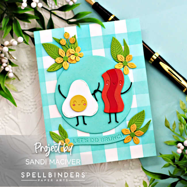 Handmade “Let’s Do Brunch” card created with the Spellbinders Small Die of the Month for November featuring a cute bacon and egg duo holding hands on a turquoise dinner plate. The card includes cheerful yellow flowers and green leaves die-cut with the Citrus Spritz Die Set, arranged around the plate. The background showcases a teal-and-white gingham pattern, ink blended with washi tape masking for the stripes. The design is bright, playful, and perfect for brunch-themed or friendship cards.