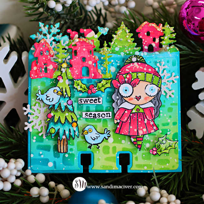 Colorful Memory Dex die card featuring AALL & Create products, with a whimsical Christmas scene on a teal and green background. The card includes a hand-colored Miss Merry character in a red dress and striped tights, pink houses along the top edge, green trees, small birds, holly leaves, snowflakes, and white paint splatter. The words “sweet season” are centered on the card, and the project is photographed among evergreen branches and bright holiday ornaments.