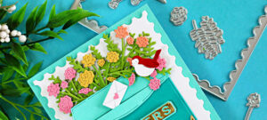 Handmade greeting card created with the Postage Box Die Set from the NEW Hero Arts My Monthly Hero Subscription program. A teal die-cut mailbox with a scalloped postage-stamp style frame is filled with colorful flowers in pink, yellow, and coral, with a tiny white envelope on top and a small bird perched on the flowers. More florals and greenery decorate the mailbox and a bundle of rolled newspapers below, all set against a teal background with coordinating metal dies arranged around the card.