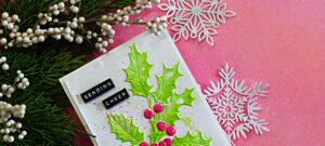 Handmade Christmas card featuring bright green embossed holly leaves with raised texture and stenciled details, accented with dimensional pink berries on a white background. Black sentiment strips read “Sending Cheer,” with paint splatter details and a festive pink backdrop.