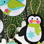 Handmade stitched felt Christmas ornaments created with new Simon Says Stamp dies, including a snowman with scarf, a teal and white bird, a black-and-white penguin with green scarf, a teal mitten with snowflake, and a holly berry sprig. Ornaments displayed against snowy bottle-brush trees and greenery.