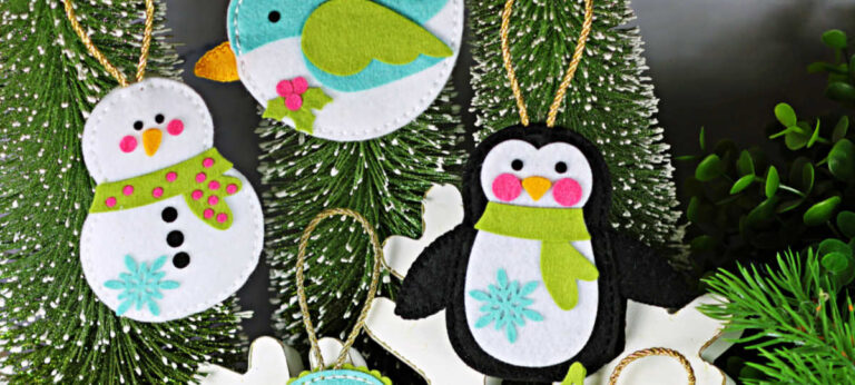 Handmade stitched felt Christmas ornaments created with new Simon Says Stamp dies, including a snowman with scarf, a teal and white bird, a black-and-white penguin with green scarf, a teal mitten with snowflake, and a holly berry sprig. Ornaments displayed against snowy bottle-brush trees and greenery.